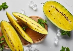 Draufsicht auf geschnittene köstliche exotische gelbe Wassermelone mit Samen auf Marmoroberfläche mit Minze, Eis und Holzschneidebrett