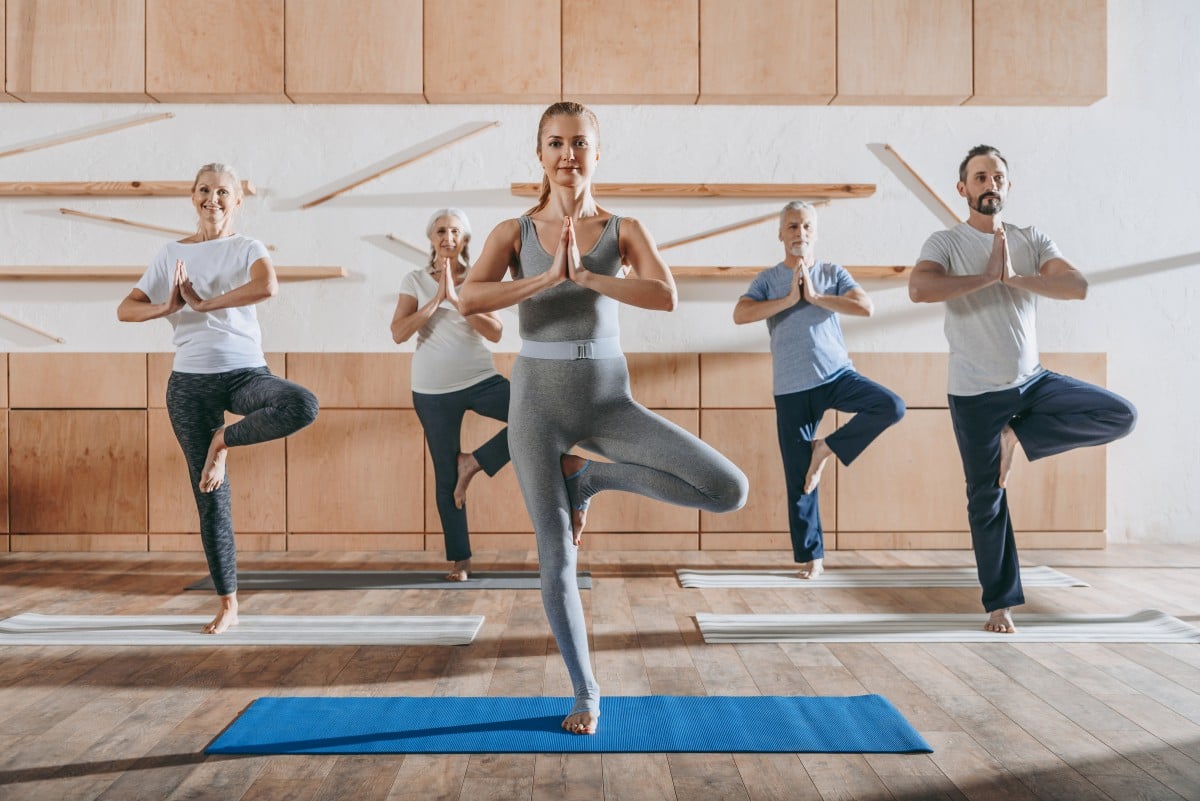 Group of senior people practicing yoga with instructor in tree pose on mats in studio