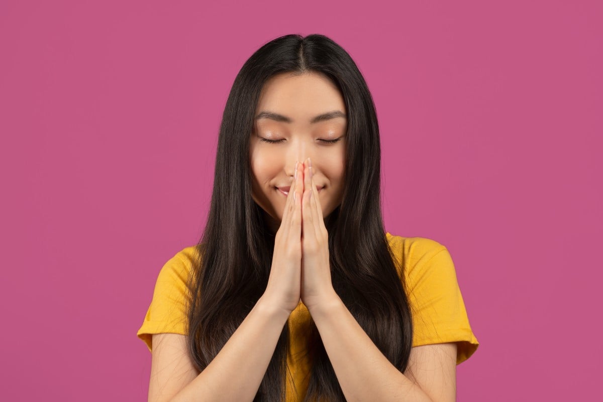 Friedliche asiatische Dame, die mit geschlossenen Augen betet, gefaltete Hände in flehender Geste hält und auf violettem Hintergrund steht