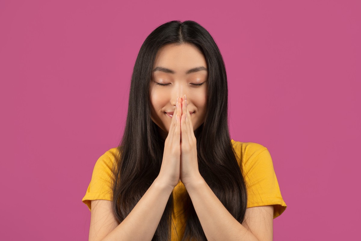 Pazful asiático orando com os olhos fechados, segurando as mãos juntas em um gesto de súplica, em pé sobre fundo violeta